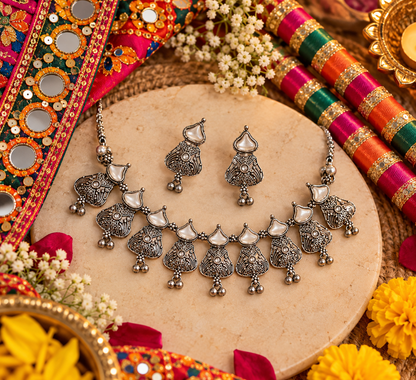 A silver-toned traditional Indian jewelry set featuring a necklace and matching earrings with ornate filigree details and white stone accents, arranged on a round beige stone plate surrounded by colorful decorative textiles, flowers, and a brass lamp.